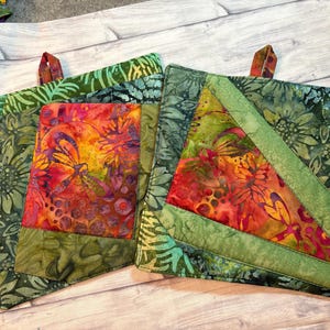 Puede incluir: Tres agarraderas acolchadas con coloridos estampados florales y de mariposas. Las agarraderas presentan tonos verdes, rojos, naranjas y amarillos. Cada agarradera tiene un lazo para colgar.