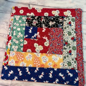 May include: A square, handmade patchwork quilt with a variety of patterned fabrics. The quilt features red, blue, yellow, and green fabrics with floral, dog, and geometric designs. The edges are neatly sewn, creating a decorative and functional item.