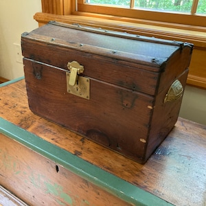 May include: A vintage wooden chest with a brass lock and handle. The chest is brown and has a worn finish. It is sitting on top of a larger wooden chest with a green painted trim.