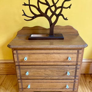 May include: A wooden dresser with three drawers and a decorative tree-shaped jewelry holder on top. The dresser has a light brown finish and the jewelry holder is dark brown.