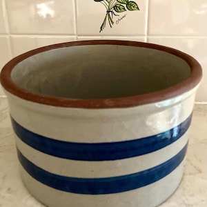 May include: A white ceramic crock with two blue stripes around the middle. The crock has a brown rim and is sitting on a white countertop.