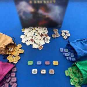 May include: A board game setup on a blue surface. The game includes a box, small rectangular game pieces, and drawstring bags in various colors: tan, purple, blue, and green. The game pieces are white with colored borders.