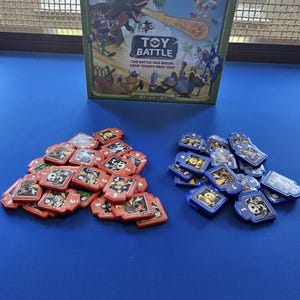 Puede incluir: Una caja de juego de mesa "Toy Battle" con el texto "The battle has begun, your troops need you!" se muestra sobre una superficie azul. Dos montones de piezas de juego, rojas y azules, están frente a la caja.