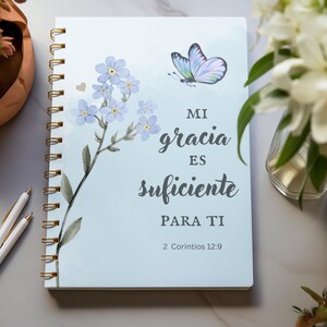 Puede incluir: Una libreta de espiral azul con una ilustración de acuarela de flores de nomeolvides y una mariposa. La libreta tiene el texto "Mi gracia es suficiente para ti" y "2 Corintios 12:9" impreso en la portada.