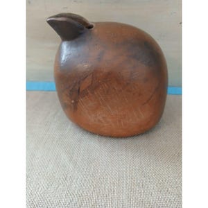 May include: A rounded, brown ceramic vessel with a smooth, polished surface and a small, protruding spout at the top left. The clay body features natural color variations, including darker patches and subtle surface markings across its exterior.