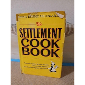 May include: A vintage cookbook titled "The Settlement Cook Book" with a bright yellow cover. The book features bold black lettering and a small illustration of a chef. The cover also includes the text "Newly Revised and Enlarged."