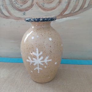 May include: A beige ceramic vase with a dark blue rim and white snowflake design. The vase has a speckled texture and a rounded shape, perfect for displaying flowers or as a decorative piece. The vase is approximately 6 inches tall.