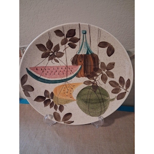 May include: Round ceramic plate with a speckled beige background. The plate features a colorful illustration of a watermelon slice, a bottle, and a green melon, with brown leaves and stems. The plate is supported by a clear plastic stand.
