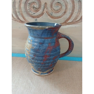 Peut inclure: Une tasse en céramique au corps bombé avec des stries horizontales et une anse texturée. L'émail présente un mélange de tons bleu profond, brun terreux et beige, rehaussé de petites empreintes circulaires marquées sur la surface.