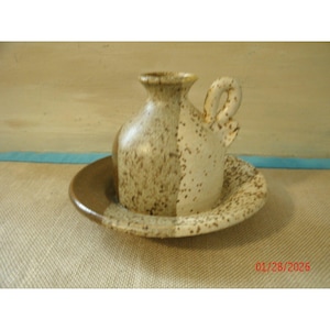 May include: A speckled ceramic creamer and saucer set. The creamer has a handle and a narrow spout. The set is in shades of brown and cream, with a textured, speckled finish. The saucer is round with a raised edge.