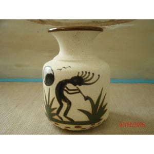 May include: A small, off-white ceramic vase with a brown rim and base. The vase features a black silhouette of a figure playing a flute, with green plant-like designs. A black circle and two birds are also depicted.