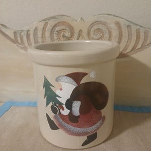 May include: A cream-colored ceramic crock with a festive Santa Claus design. Santa is depicted carrying a green Christmas tree and a brown sack. The crock has a cylindrical shape and is suitable for kitchen or holiday decor.