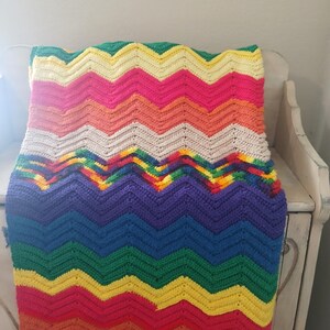 May include: A colorful, handmade crochet blanket with a chevron pattern. The blanket features a vibrant array of colors, including red, yellow, green, blue, purple, and pink. The blanket is draped over a white wooden surface.