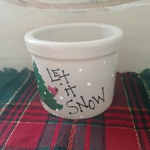 May include: A small, beige ceramic pot with a rounded shape. The pot is decorated with a green Christmas tree, a red bird, and the words "Let it Snow" in black lettering. White dots represent snowflakes.