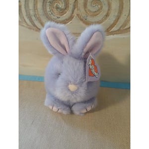 May include: A plush, lavender-colored bunny rabbit with pink inner ears and a white nose. The stuffed animal has a tag that reads "Puffkins" in a heart-shaped design. The bunny is sitting on a neutral-colored surface.
