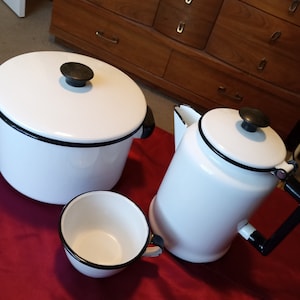 Peut inclure: Un ensemble de trois articles de cuisine en émail blanc avec une bordure noire. L'ensemble comprend une grande casserole avec un couvercle, un pichet plus petit et une petite tasse.