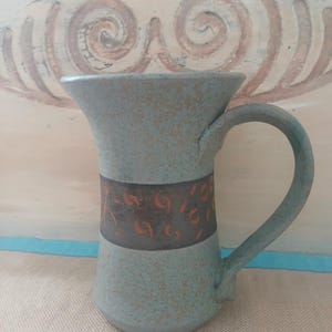 Peut inclure: Une tasse en céramique faite à la main avec un glaçage bleu clair moucheté. Une bande foncée avec un motif brun rougeâtre entoure le milieu. La tasse a une anse incurvée et un dessus évasé.