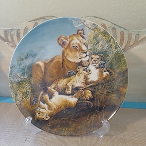 May include: A decorative plate depicting a lioness and her cubs in a naturalistic scene. The plate has a blue sky background and a detailed illustration of the lions in shades of brown and tan. The plate is displayed on a clear plastic stand.