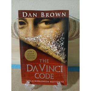May include: A hardcover book titled "The Da Vinci Code" by Dan Brown. The cover features a close-up of a face with overlaid text and the words "The Da Vinci Code" in large letters. A gold sticker reads "Now a Major Motion Picture."