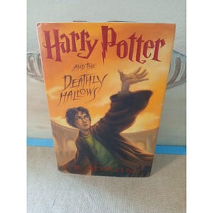 May include: A hardcover book titled Harry Potter and the Deathly Hallows by J.K. Rowling. The cover features an illustration of a character with dark hair and glasses wearing a brown robe, reaching upward against an orange and yellow sky background.