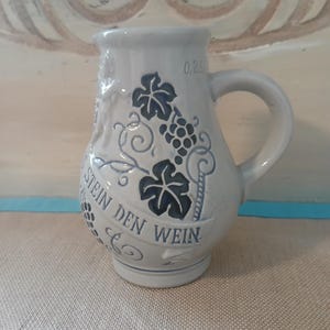 May include: A light blue ceramic pitcher with a handle, featuring a grape and leaf design in dark blue. The words "STEIN DEN WEIN" are written on the front. The pitcher has a capacity marking of 0.25L.