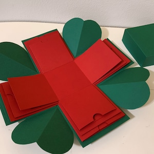 Puede incluir: Una caja explosiva hecha a mano, abierta para revelar capas de papel rojo y verde. La caja tiene forma de flor, con pétalos verdes en forma de corazón. En el interior, capas rojas con bolsillos. También se ve una caja verde separada.