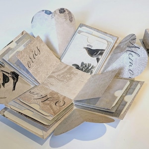 Può includere: Una scatola esplosiva beige aperta con alette a forma di cuore e una scatola rettangolare abbinata. La scatola è decorata con illustrazioni floreali e di uccelli, e le parole "Memories" e "Mem" sono scritte sui lembi. La scatola contiene piccole tasche.