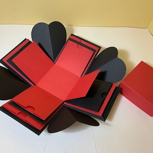 Puede incluir: Una caja de explosión roja y negra con una tapa en forma de corazón. La caja está abierta y el interior es visible. Hay una caja roja más pequeña al lado de la caja de explosión.