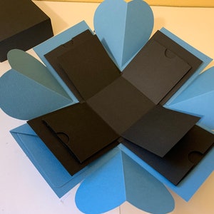 May include: A blue and black explosion box with multiple flaps that open to reveal smaller compartments. The box is made of cardstock and has a heart-shaped design.