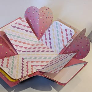 Puede incluir: Una caja explosiva hecha a mano, abierta para revelar papel estampado y solapas en forma de corazón. La caja es rosa con confeti y paneles interiores estampados. Una pequeña caja estampada es visible en el fondo.