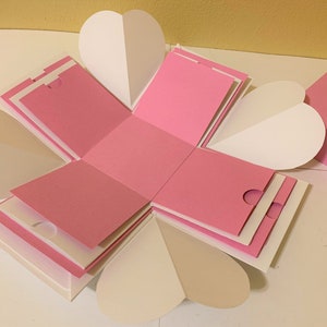 以下が含まれることがあります： ハート型のフラップが付いたピンクと白の爆発ボックス。ボックスが開いており、フラップが後ろに折り畳まれ、内部が明らかになっています。ボックスは厚紙製で、質感のある仕上げになっています。
