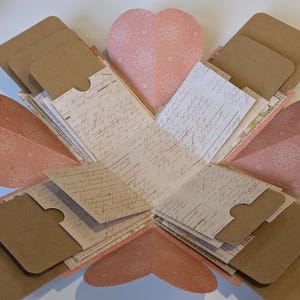 May include: An open, heart-shaped explosion box with multiple layers. The box features pink heart-shaped panels with a floral pattern, and brown cardstock pockets. Inside are pages with handwritten script.