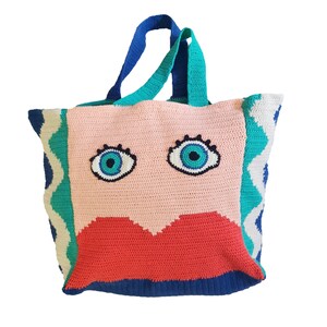 May include: A crocheted tote bag with a pink face, blue eyes, and a red mouth. The bag has a blue, green, and white chevron pattern on the sides.