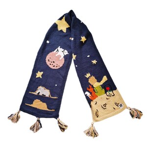 Puede incluir: Una bufanda de punto azul con un diseño caprichoso que presenta al Principito, un zorro y una rosa. La bufanda tiene borlas en cada extremo y está decorada con estrellas y un planeta.