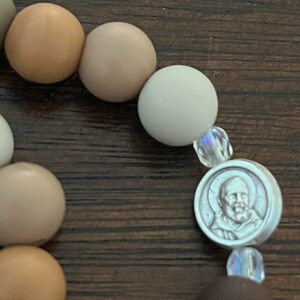 May include: A brown, beige and white beaded bracelet with a silver charm featuring a man's face. The charm is surrounded by clear crystal beads.