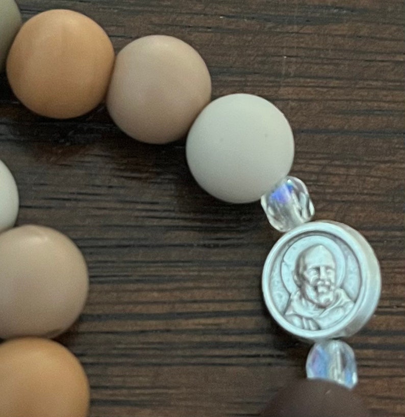 May include: A brown, beige and white beaded bracelet with a silver charm featuring a man's face. The charm is surrounded by clear crystal beads.