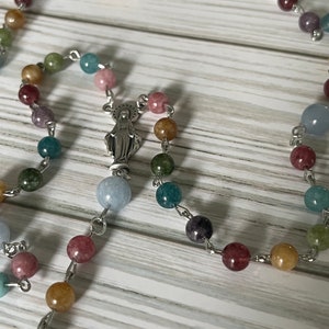 May include: A colorful beaded rosary with a silver charm of the Virgin Mary. The beads are made of various semi-precious stones, including blue, green, pink, and yellow.