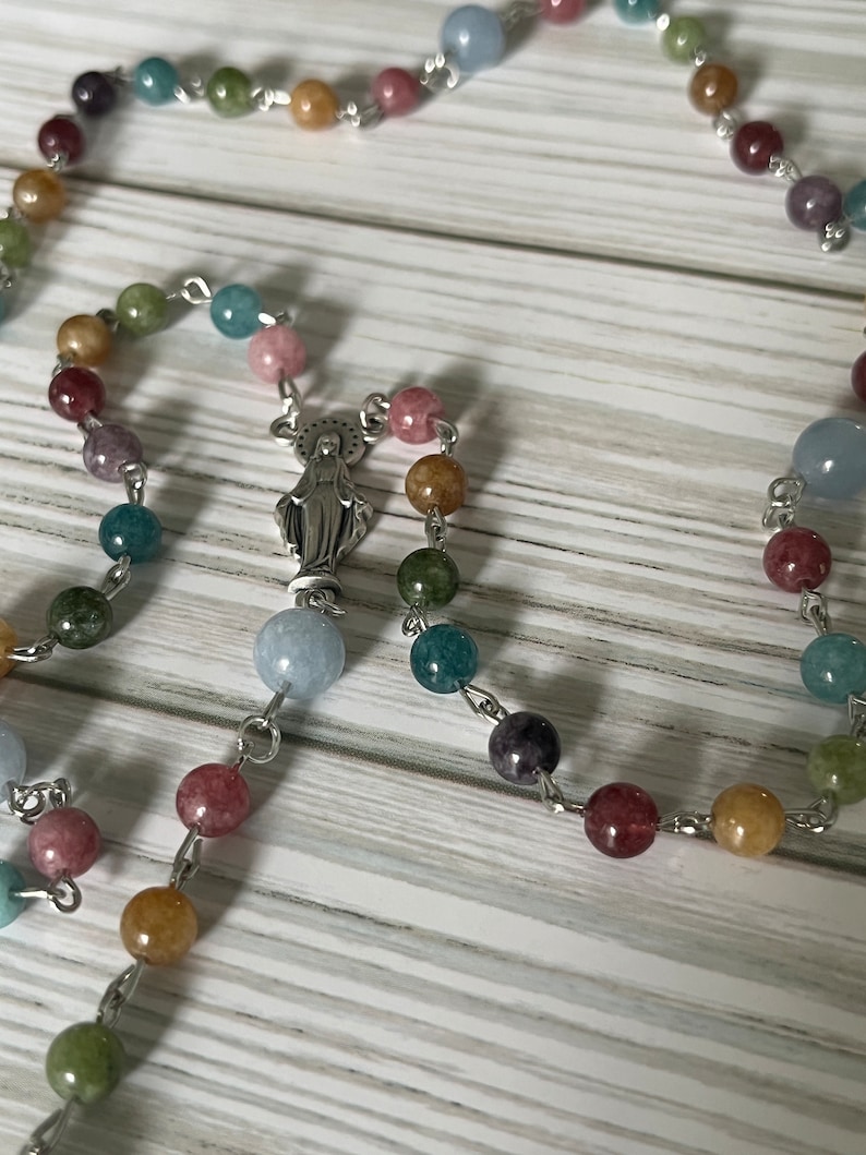May include: A colorful beaded rosary with a silver charm of the Virgin Mary. The beads are made of various semi-precious stones, including blue, green, pink, and yellow.