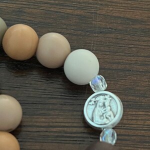 May include: A close-up of a beaded bracelet with a silver charm featuring a religious image. The beads are various shades of brown and white.