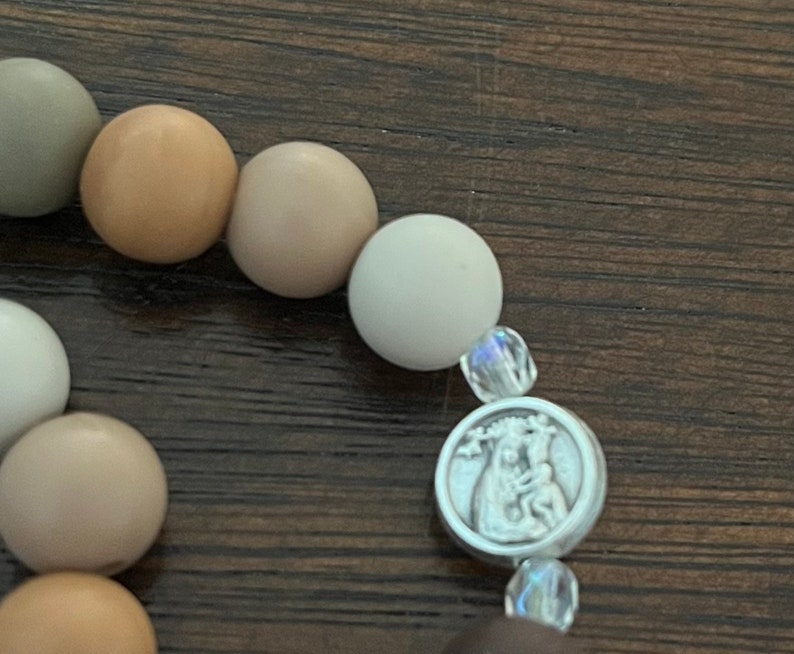 May include: A close-up of a beaded bracelet with a silver charm featuring a religious image. The beads are various shades of brown and white.