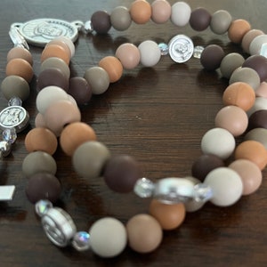 May include: A brown, beige and grey beaded rosary with silver accents. The beads are made of silicone and are strung on a silver chain. The rosary has a silver crucifix and a silver medal of the Virgin Mary.