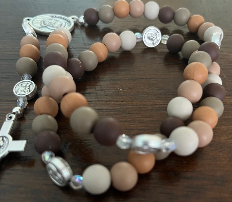 May include: A brown, beige and grey beaded rosary with silver accents. The beads are made of silicone and are strung on a silver chain. The rosary has a silver crucifix and a silver medal of the Virgin Mary.