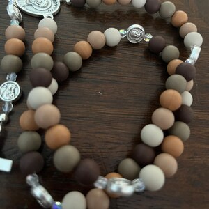 May include: A brown, beige and grey beaded rosary with a silver charm featuring a religious figure. The beads are made of wood and have a matte finish.