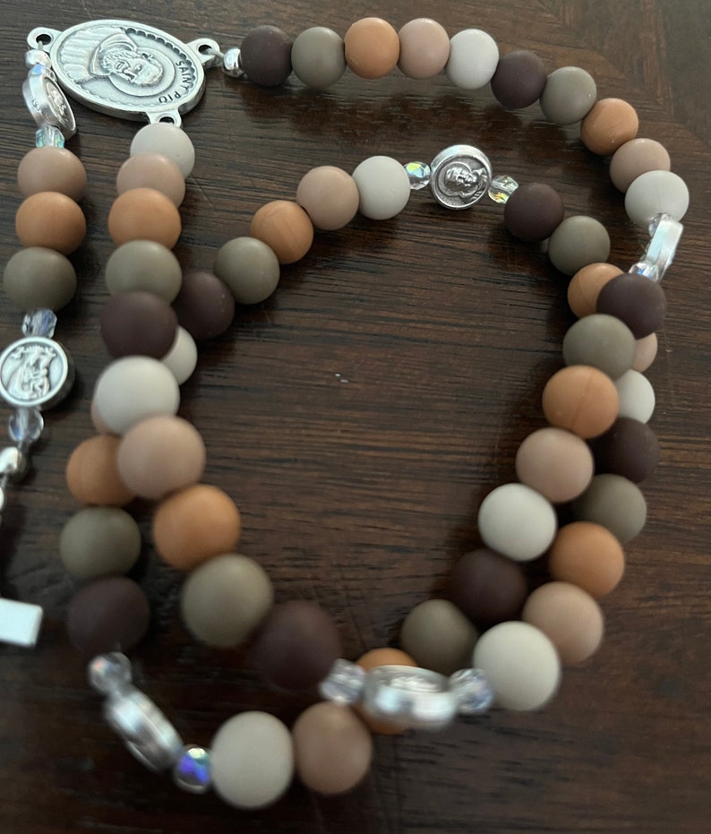 May include: A brown, beige and grey beaded rosary with a silver charm featuring a religious figure. The beads are made of wood and have a matte finish.