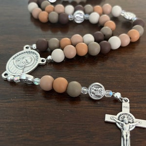 May include: A brown, beige and grey rosary with a silver crucifix and a silver medal of Saint Pio. The rosary beads are made of silicone.