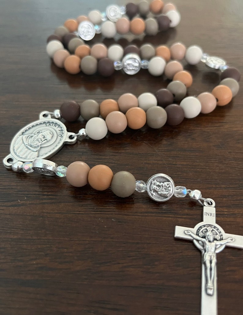 May include: A brown, beige and grey rosary with a silver crucifix and a silver medal of Saint Pio. The rosary beads are made of silicone.