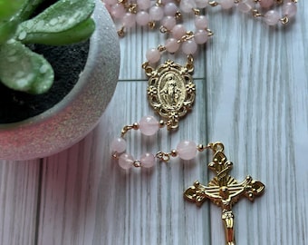 May include: A rosary with pink gemstone beads and gold-colored metal accents. The rosary features a gold-colored crucifix and a medallion with a religious figure. A small potted succulent is in the background.