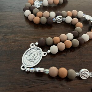 May include: A brown, beige and grey beaded rosary with a silver charm featuring a picture of Saint Pio. The beads are made of a soft, pliable material.