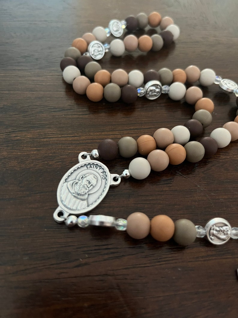 May include: A brown, beige and grey beaded rosary with a silver charm featuring a picture of Saint Pio. The beads are made of a soft, pliable material.