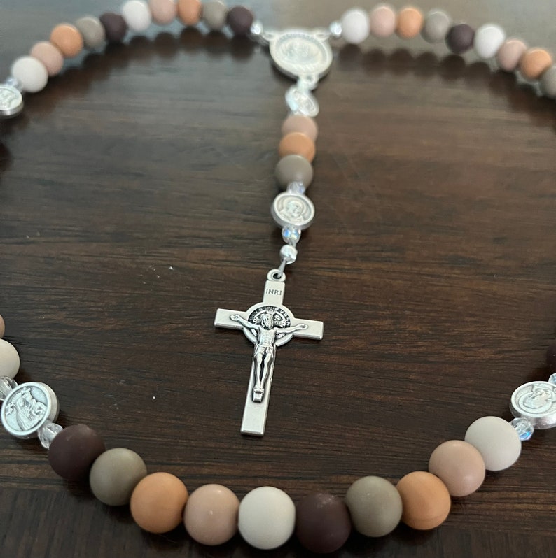 May include: A brown, beige and white beaded rosary with a silver crucifix and a silver charm featuring the image of Saint Francis of Assisi. The rosary is laid out on a wooden surface.